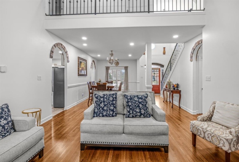Spacious living area featuring hardwood flooring, recessed lighting, white wall paneling, brick archways, and an upper-level balcony with iron railings