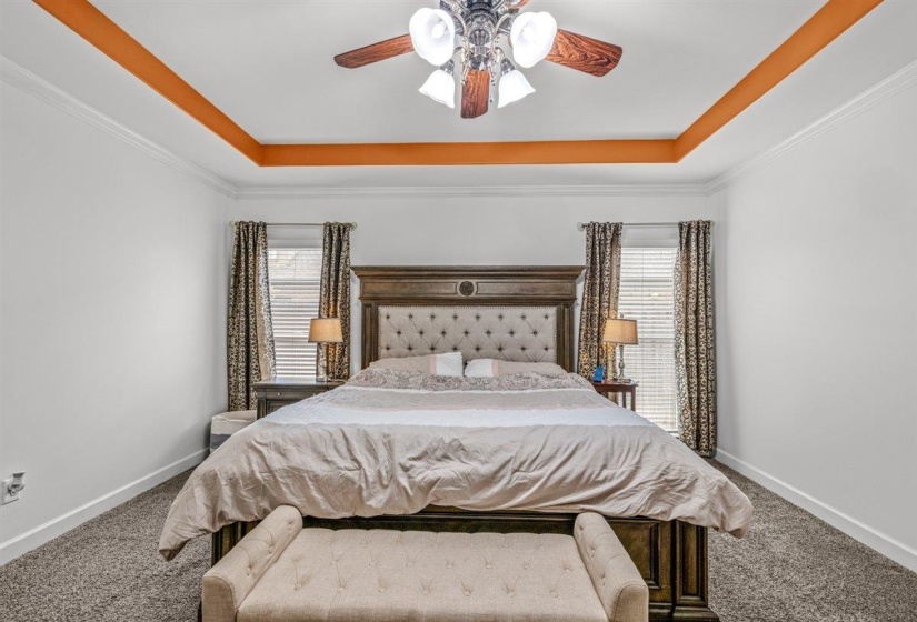 Tray ceiling with crown molding and recessed orange accents, overhead ceiling fan with light fixture, and two windows with blinds and patterned drapes