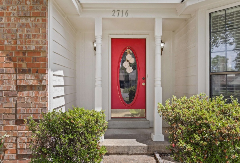 Vibrant red entry door with oval glass insert, flanked by white decorative columns and wall-mounted light fixtures