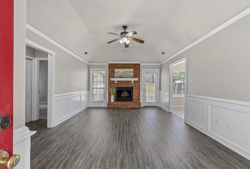 Spacious living area featuring wood-finish flooring, a vaulted ceiling with a ceiling fan, wainscoting, and a brick fireplace with a white mantel