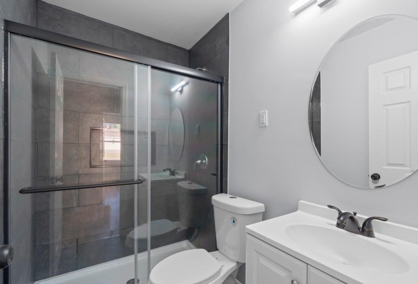 Bathroom featuring a glass-enclosed shower with dark gray tile surround, a white vanity with integrated sink, a circular mirror, and a modern light fixture