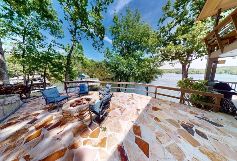 Expansive flagstone patio featuring a built-in stone fire pit, wood railing, and mature trees