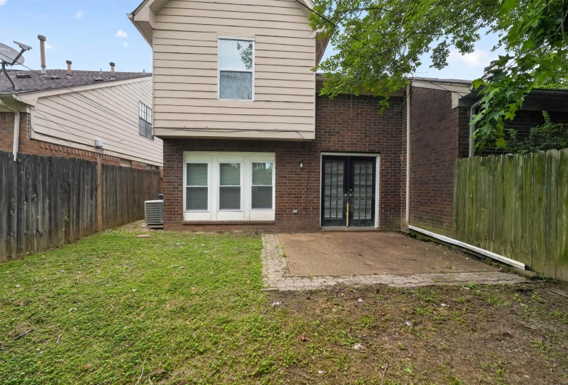 Rear exterior with a brick and siding facade, featuring a ground-level patio with an adjacent grassy area, a privacy fence, and a double-door entryway