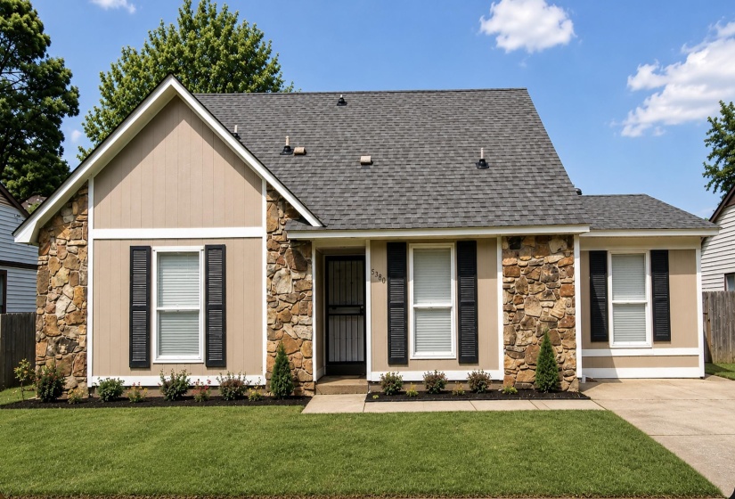 Exterior featuring stone façade accents, beige siding, black louvered shutters, a dark shingle roof, and a meticulously maintained front lawn with landscaping