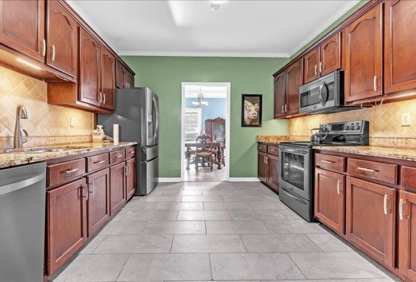 Galley kitchen featuring wood-finish cabinetry, stone countertops, stainless steel appliances, a tiled backsplash, and light-toned tile flooring