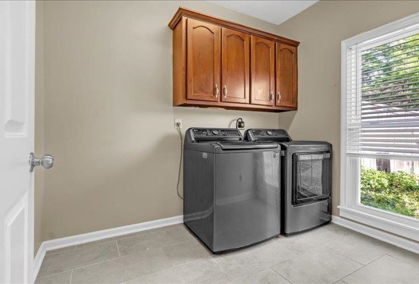Utility area featuring light neutral wall paint, wood cabinetry with gold-toned hardware, a white-framed window with blinds, and rectangular floor tiling