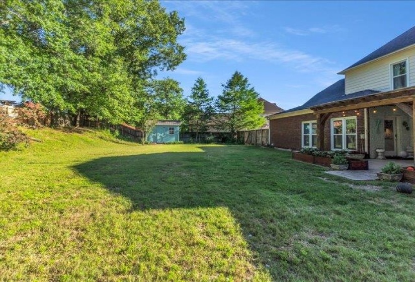 Expansive backyard featuring a large grassy area, mature trees, a detached storage shed, a brick exterior on the lower level, and a covered patio with a wooden pergola