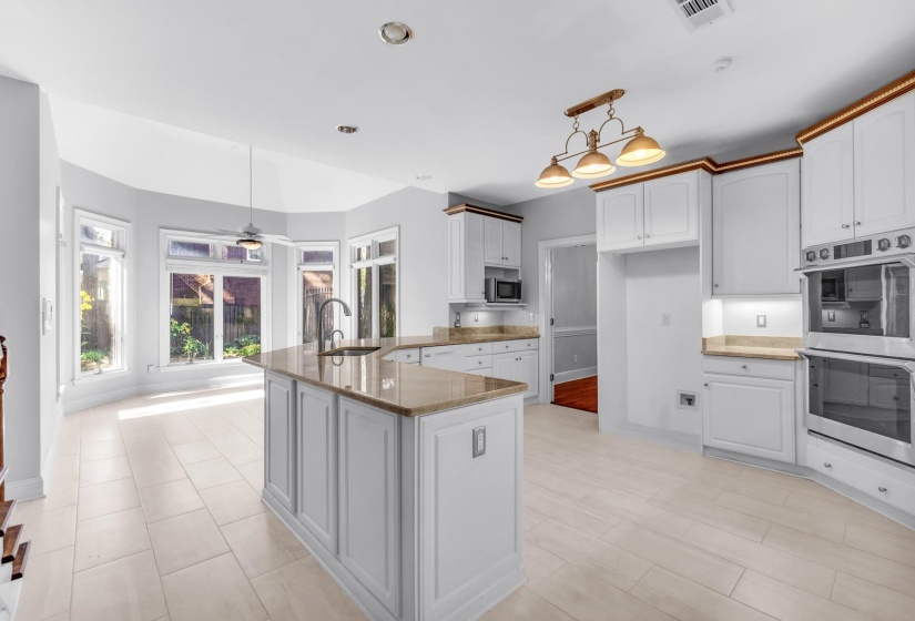 Open-concept kitchen and dining area featuring a central island with sink, extensive white cabinetry with crown molding, stainless steel built-in double ovens, and a bay window configuration