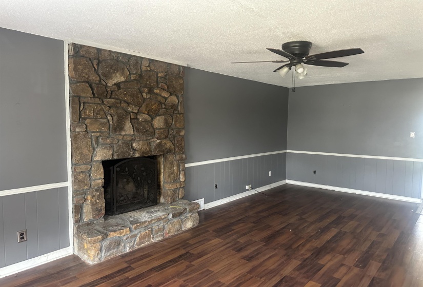Spacious living area featuring a stone-clad fireplace with an ornate screen, wood-finish flooring, grey painted walls, and white wainscoting