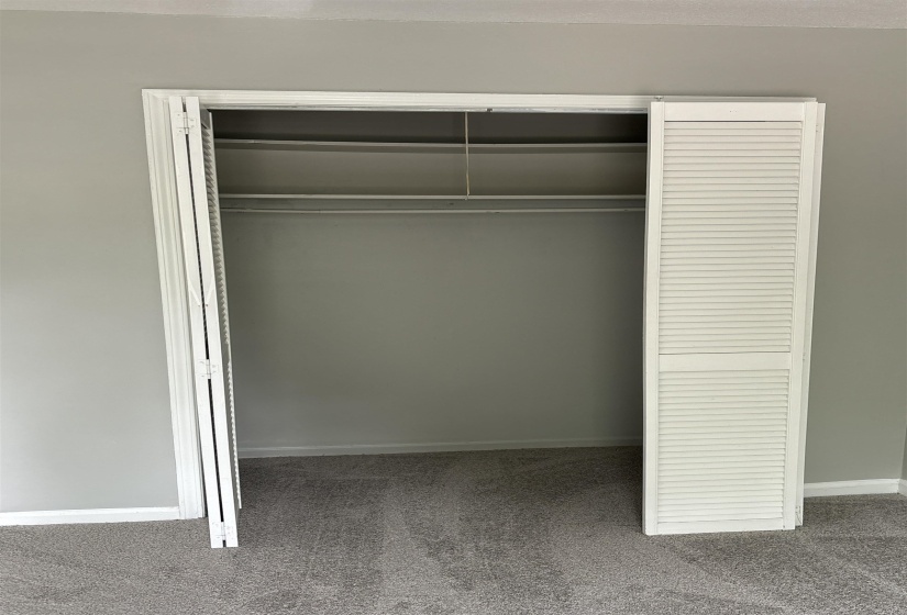 Recessed closet with white louvered bifold doors, integrated shelving, and hanging rod, complemented by grey wall paint and grey carpet flooring