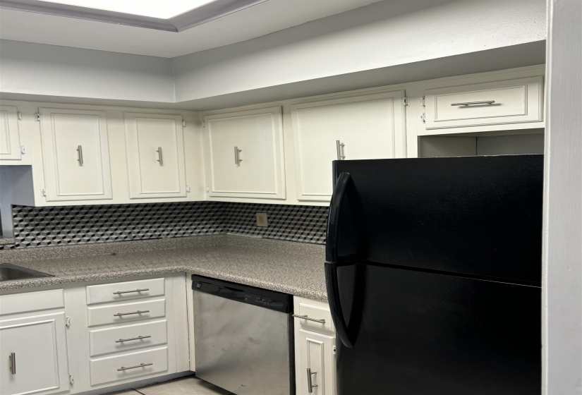 Kitchen featuring extensive white cabinetry, a black refrigerator, stainless steel dishwasher, speckled countertops, and a geometric pattern backsplash