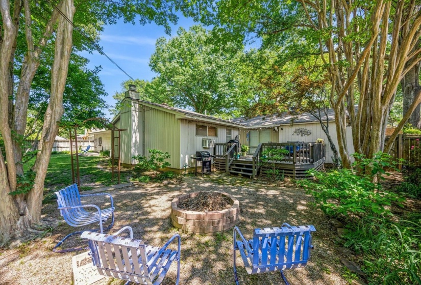 Spacious backyard featuring mature trees, a wooden deck with railing, a brick fire pit, and a shingle roofline
