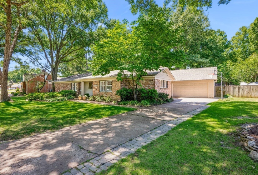 Brick facade home featuring a two-car garage, mature trees, a paved driveway with paver accents, and a manicured front lawn