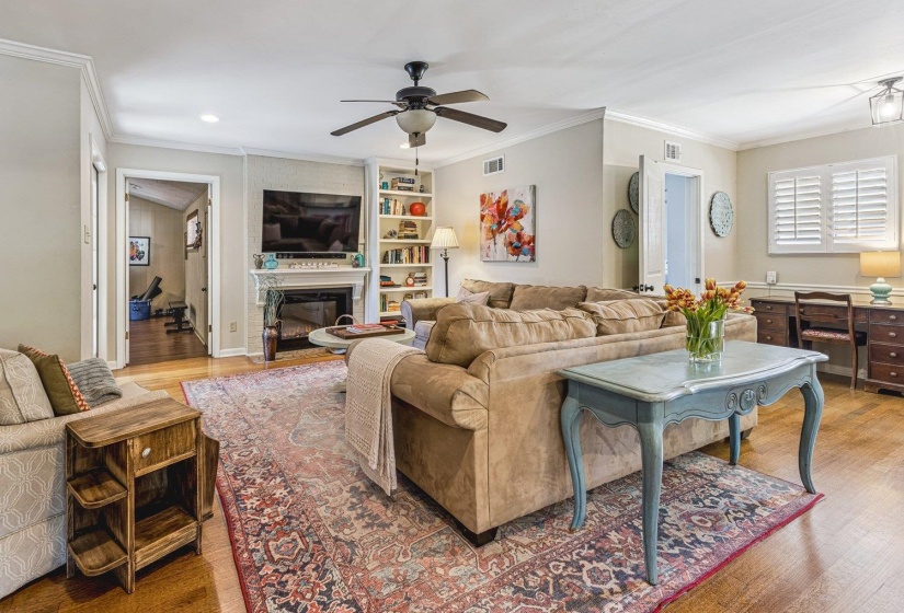 Living area featuring wood-finish flooring, a ceiling fan, a fireplace with a mantel, built-in shelving, and plantation shutters