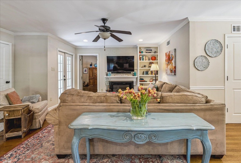 Living area featuring hardwood flooring, crown molding, a ceiling fan, built-in shelving, and a fireplace with mantle