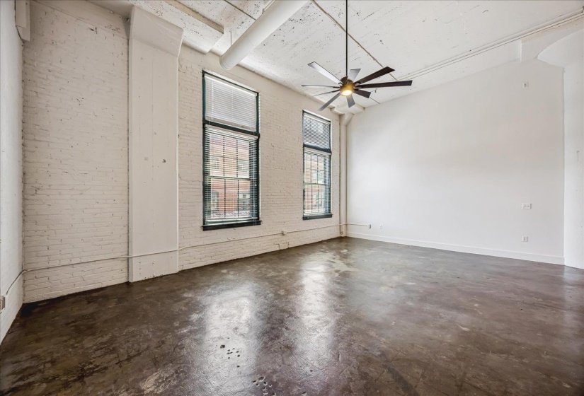 Spacious room featuring exposed brick walls, a high ceiling, two tall windows with blinds, a ceiling fan, and concrete flooring