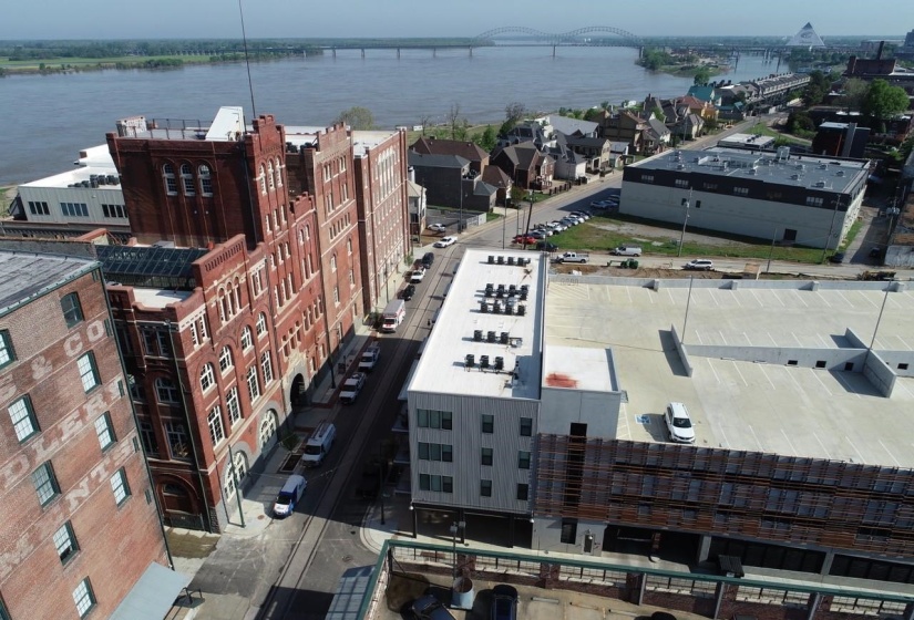 Aerial perspective showcasing a mixed-use urban landscape featuring brick and modern buildings, a multi-level parking structure, and a prominent river with a bridge in the background