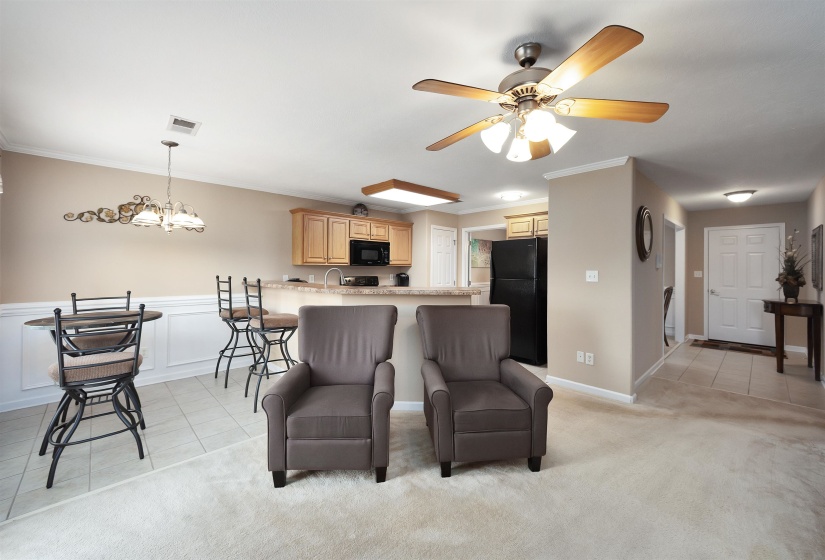 Open-concept living space featuring a ceiling fan with light kit, light-toned carpeting, and a kitchen with wood cabinetry, black appliances, and a breakfast bar