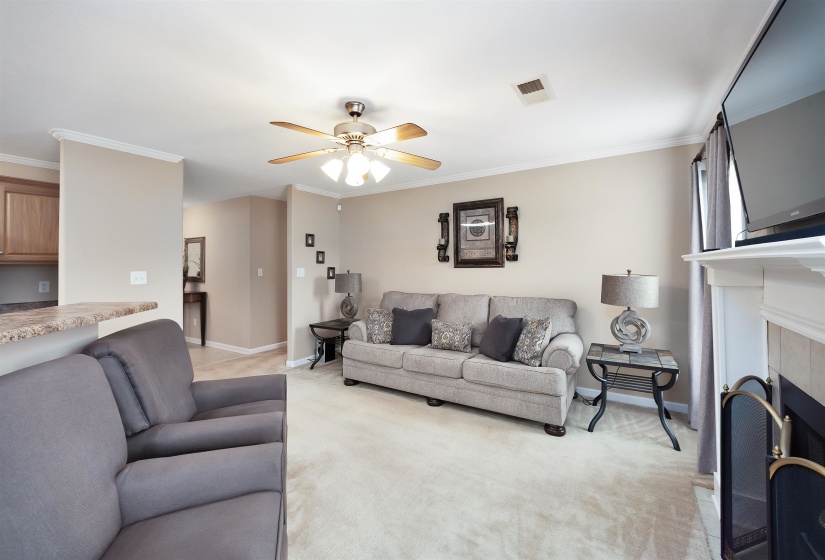 Open-concept living area with neutral carpeting, a ceiling fan, crown molding, and a built-in fireplace featuring a white mantel