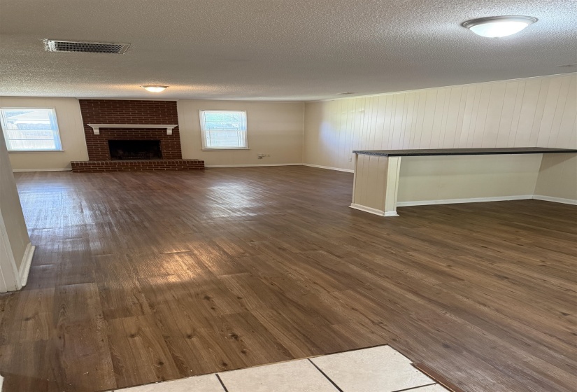 Spacious living area featuring wood-finish flooring, a brick fireplace with a white mantel, multiple windows, recessed lighting, and a built-in breakfast bar with a dark countertop