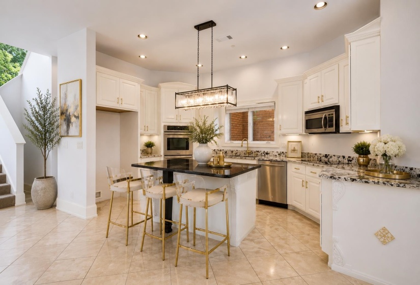 Open-concept kitchen featuring an island with a dark countertop, white cabinetry with gold-tone hardware, stainless steel appliances, and light-toned tile flooring