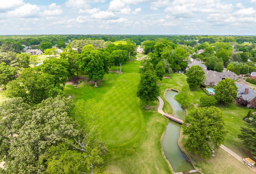 Expansive green space featuring a golf course fairway, mature trees, a meandering stream, and a footbridge