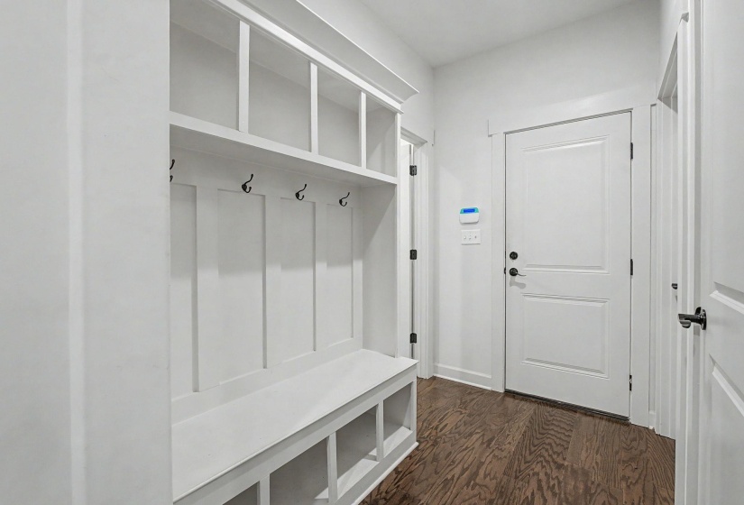 Built-in white mudroom bench featuring upper shelving, coat hooks, and lower cubbies, complemented by dark wood-finish flooring and white paneled doors
