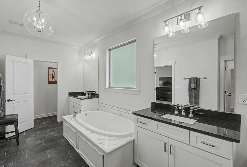 Bathroom featuring a built-in tub with a marble-look surround, dual vanity areas with white cabinetry and dark countertops, a large framed mirror, and rectangular floor tiling