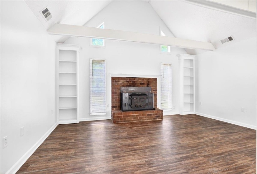 Vaulted ceiling featuring exposed beams, a brick fireplace with a wood-burning stove insert, wood-finish flooring, and built-in shelving