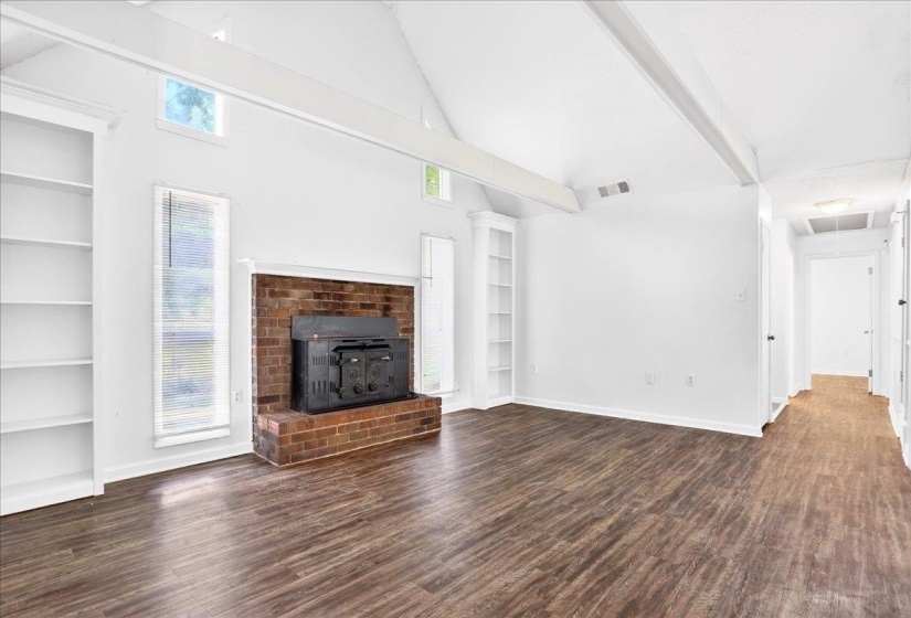 Vaulted ceiling with exposed beam detailing, wood-finish flooring, brick fireplace with metal insert, built-in shelving, and transom windows
