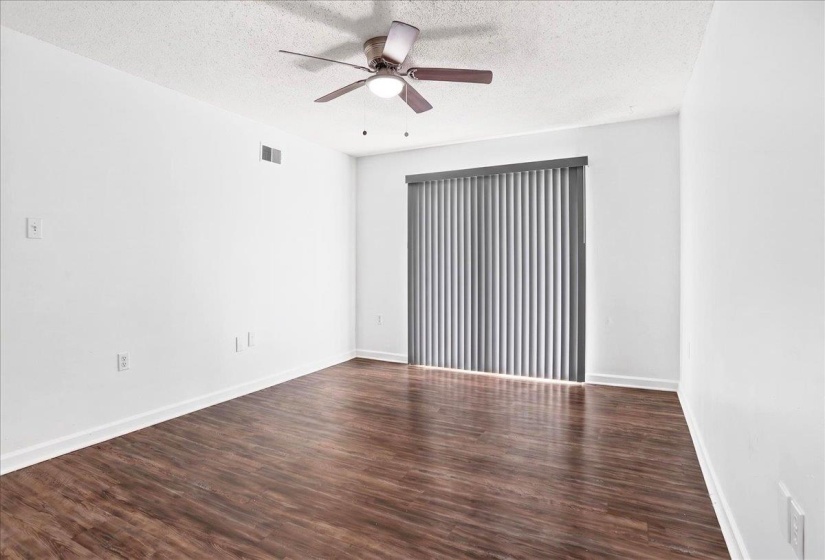 Spacious room featuring wood-finish flooring, a ceiling fan with integrated lighting, and a vertical blind-equipped sliding glass door