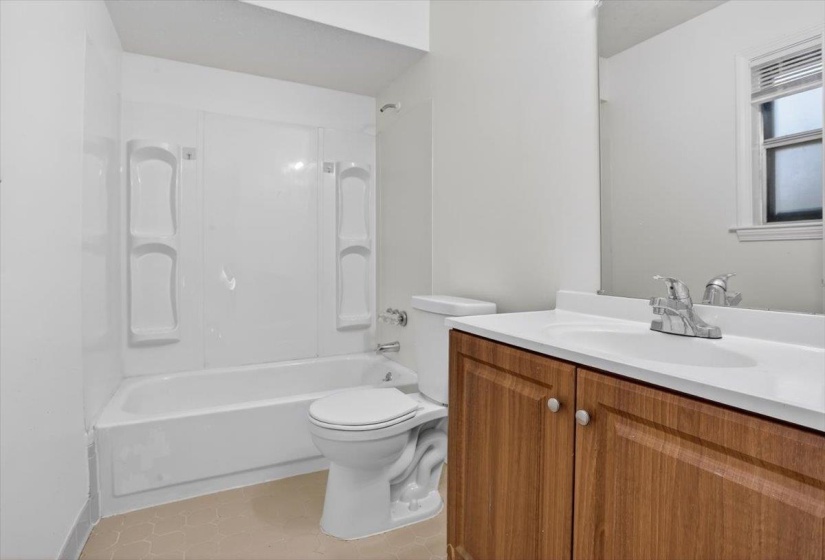 Bathroom featuring a vanity with wood-finish cabinetry and a white countertop, a mirror, a toilet, and a white tub with a white surround