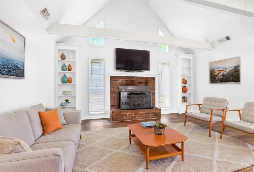 Living area featuring a brick fireplace with a wood-burning insert, vaulted ceilings with exposed beams, built-in shelving, windows, and wood-finish flooring (staged)
