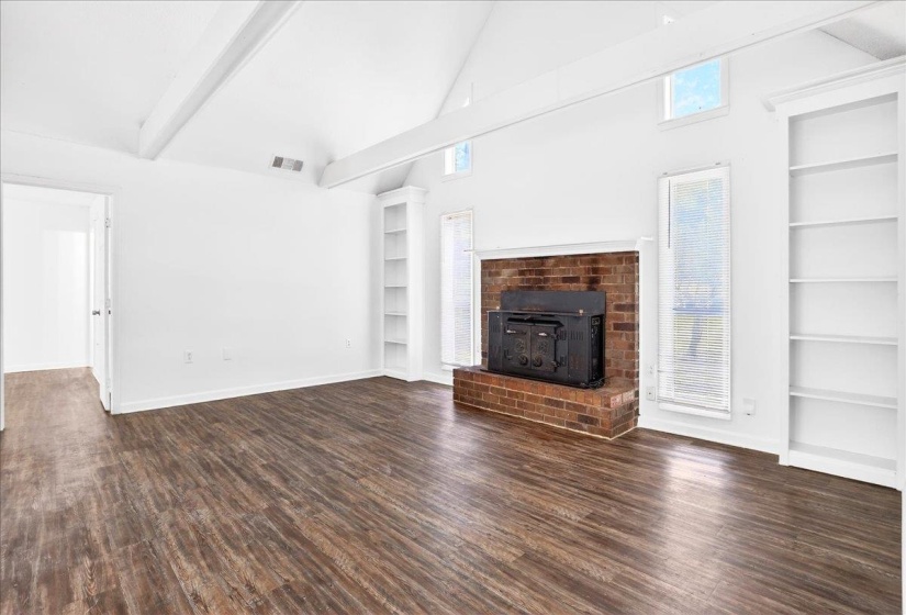 Vaulted ceiling with exposed beams, wood-finish flooring, brick fireplace with a built-in insert, built-in shelving, and large windows with blinds