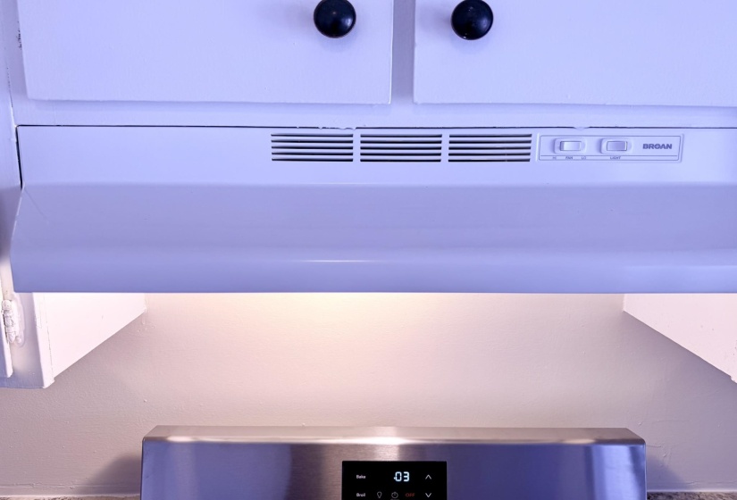 White range hood with integrated lighting, positioned below white cabinetry with black round knobs, above a stainless steel range with a digital display