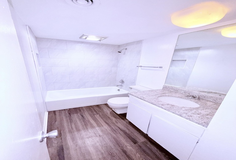 Bathroom featuring wood-finish flooring, a white vanity with a speckled countertop, a large wall-mounted mirror, and a tiled shower-tub combination with a built-in niche