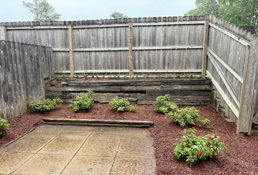 Patio area featuring concrete pavers, a privacy fence, and a terraced garden bed with wood retaining timbers, red mulch, and established plantings