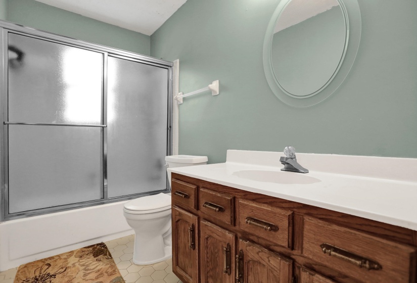 Bathroom featuring a wood-finish vanity with a white countertop and integrated sink, an oval wall-mounted mirror, and a bathtub with sliding frosted glass doors