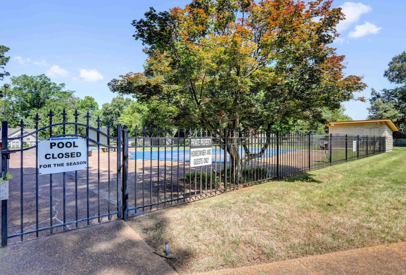 Community pool area featuring a black wrought iron fence, a pool house with horizontal siding, and mature trees providing natural shade