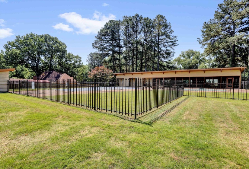 Fenced pool area with a surrounding grassy expanse, featuring a modern building with floor-to-ceiling windows and an adjacent storage structure