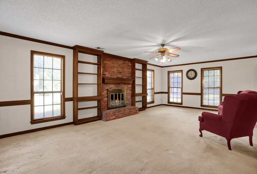 Spacious living area featuring a brick fireplace with a wood mantel, built-in shelving, multiple windows, and a ceiling fan