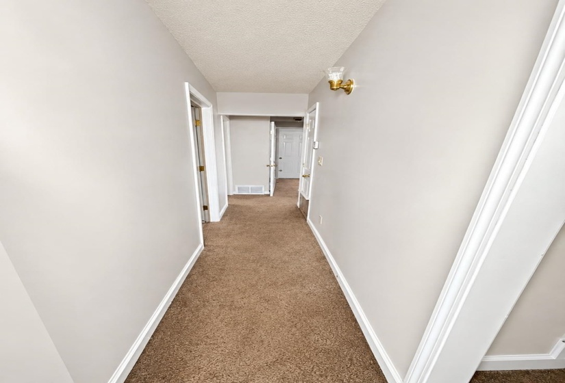 Hallway featuring light-toned walls, brown carpet flooring, white baseboards, and a gold-toned wall sconce with a glass shade AI-Enhanced Photo — May Not Reflect Exact Current Condition