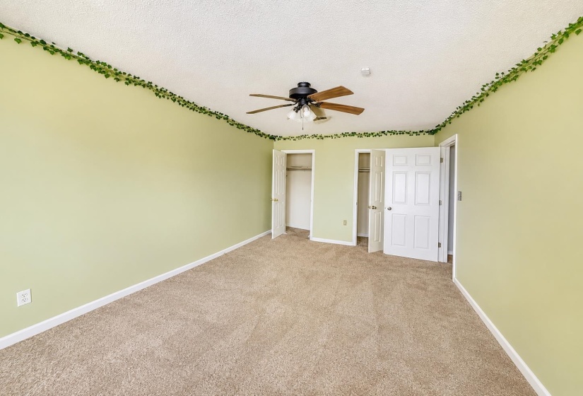 Carpeted room featuring a ceiling fan with light fixture, two white paneled doors, and two open closets with shelving AI-Enhanced Photo — May Not Reflect Exact Current Condition