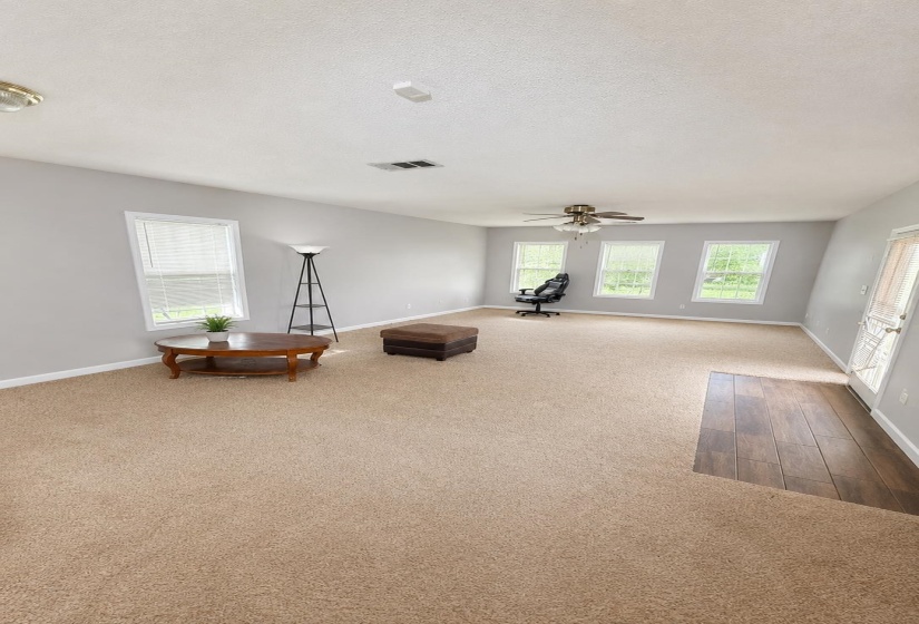 Spacious room featuring neutral carpeting, multiple windows for natural illumination, a ceiling fan, light gray painted walls, and wood-finish flooring at the entrywayAI-Enhanced Photo — May Not Reflect Exact Current Condition