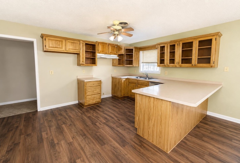 Kitchen featuring wood-finish flooring, a ceiling fan, light wood cabinetry, a corner sink with a window, and a peninsula countertop AI-Enhanced Photo — May Not Reflect Exact Current Condition