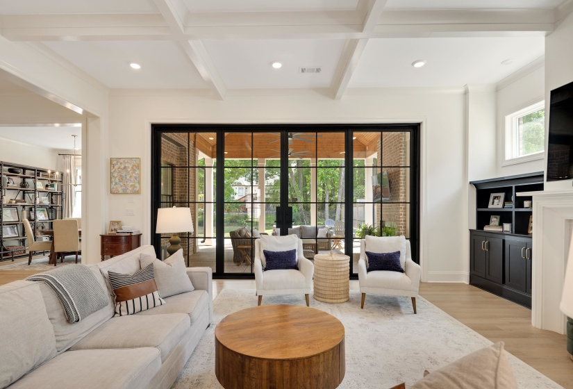 Expansive living area with coffered ceilings, large black-framed glass doors, light wood-finish flooring, recessed lighting, and built-in cabinetry