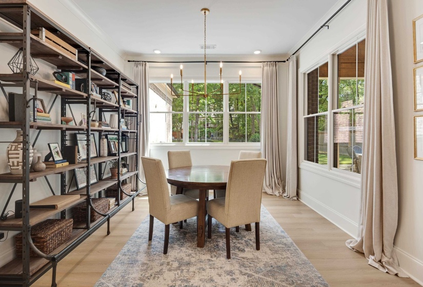 Dining area featuring light wood-finish flooring, an extensive built-in shelving system with dark metal framing and wood shelves, large multi-pane windows with white trim, and a contemporary linear chandelier with brass accents