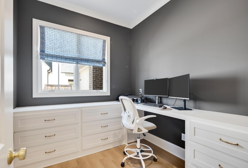 Dedicated workspace featuring built-in white cabinetry with gold-tone hardware, a generous built-in desk surface, and wood-finish flooring