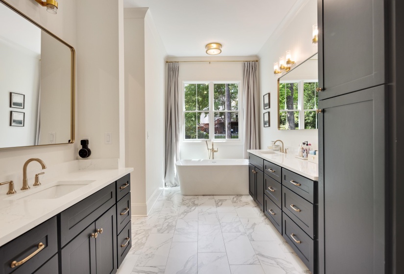 Freestanding soaking tub with a floor-mounted brass faucet, flanked by two dark gray vanity cabinets featuring white countertops and brass hardware