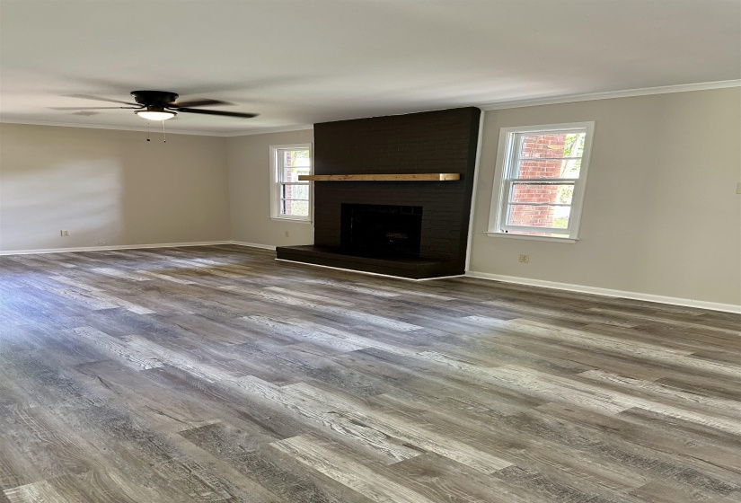 Spacious living area featuring wide plank wood-finish flooring, a dark brick fireplace with a natural wood mantel, two large windows, and a ceiling fan with integrated lighting